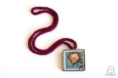 Kwadratowy wisiorek ze szkła ze starym znaczkiem pocztowym ilustrującym kwiat. Naszyjnik na bordowym łańcuszku.